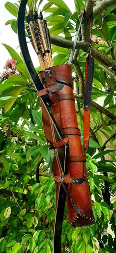 a leather archery quiver hangs from a tree branch, holding a recurve bow and several arrows. the quiver is crafted from rich brown leather with decorative stitching and multiple compartments. the bow is also made of wood, with a dark finish. the background is a lush green foliage, suggesting a sunny outdoor setting.