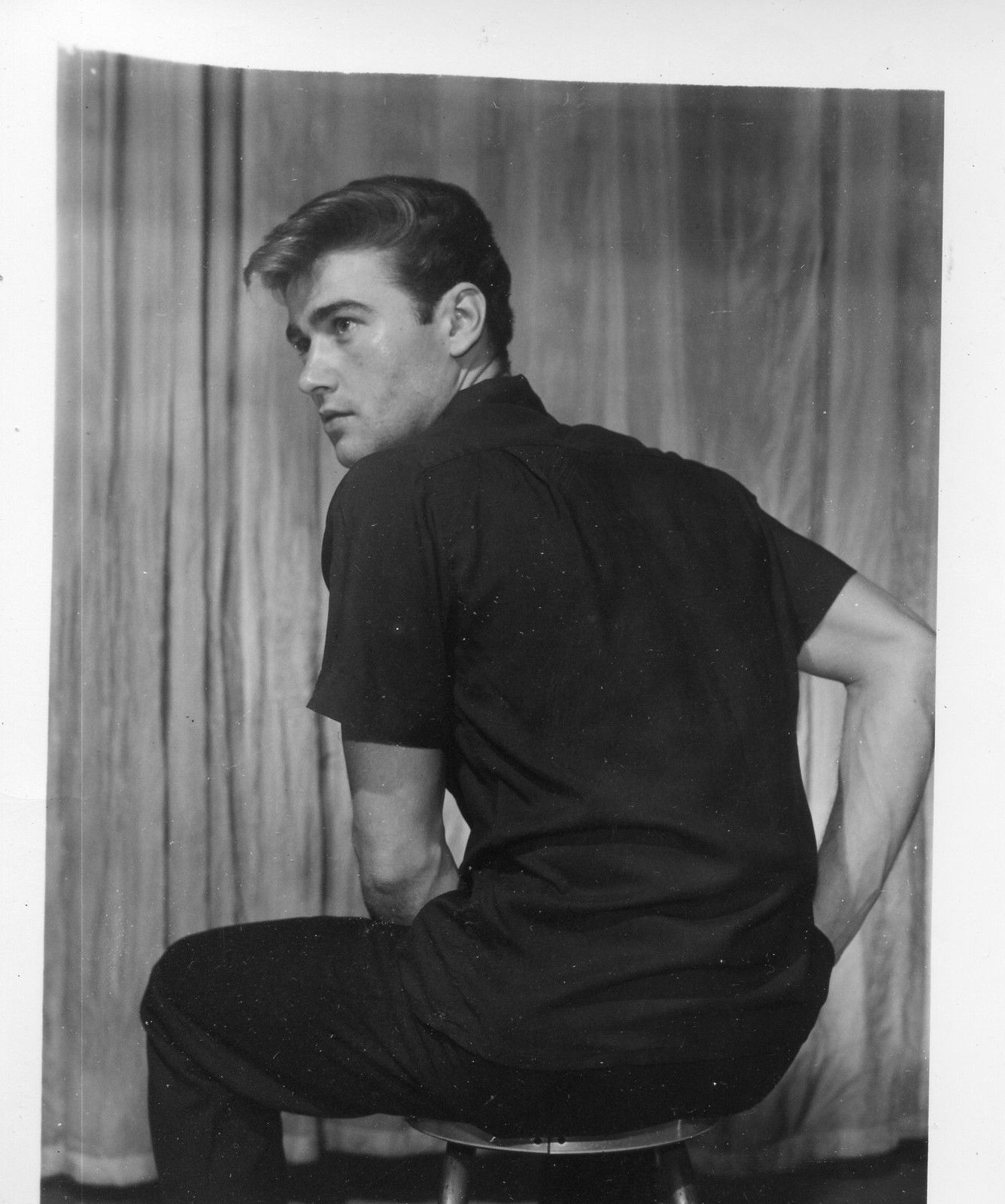 a black and white portrait of a young clint eastwood. he is seated on a stool, wearing a dark shortsleeved shirt and dark pants. he is turned slightly to the side, looking over his left shoulder, with a neutral expression on his face. the background is a plain, textured curtain. the lighting is soft and even, typical of studio portraiture.