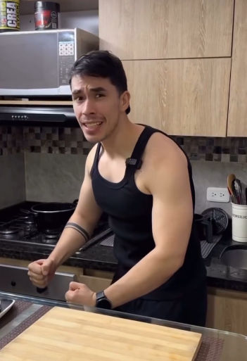 a man with a muscular build is standing in a kitchen, appearing excited and about to cook. he is looking directly at the camera with a wideeyed expression and clenching his fists in front of him. he is wearing a black tank top and a black baseball cap. the kitchen features wooden cabinets, a black countertop, and stainless steel appliances. a cutting board and a waffle iron are visible on the counter. the overall lighting suggests indoor artificial lighting. the perspective is from eye level, giving the impression of a selfie.