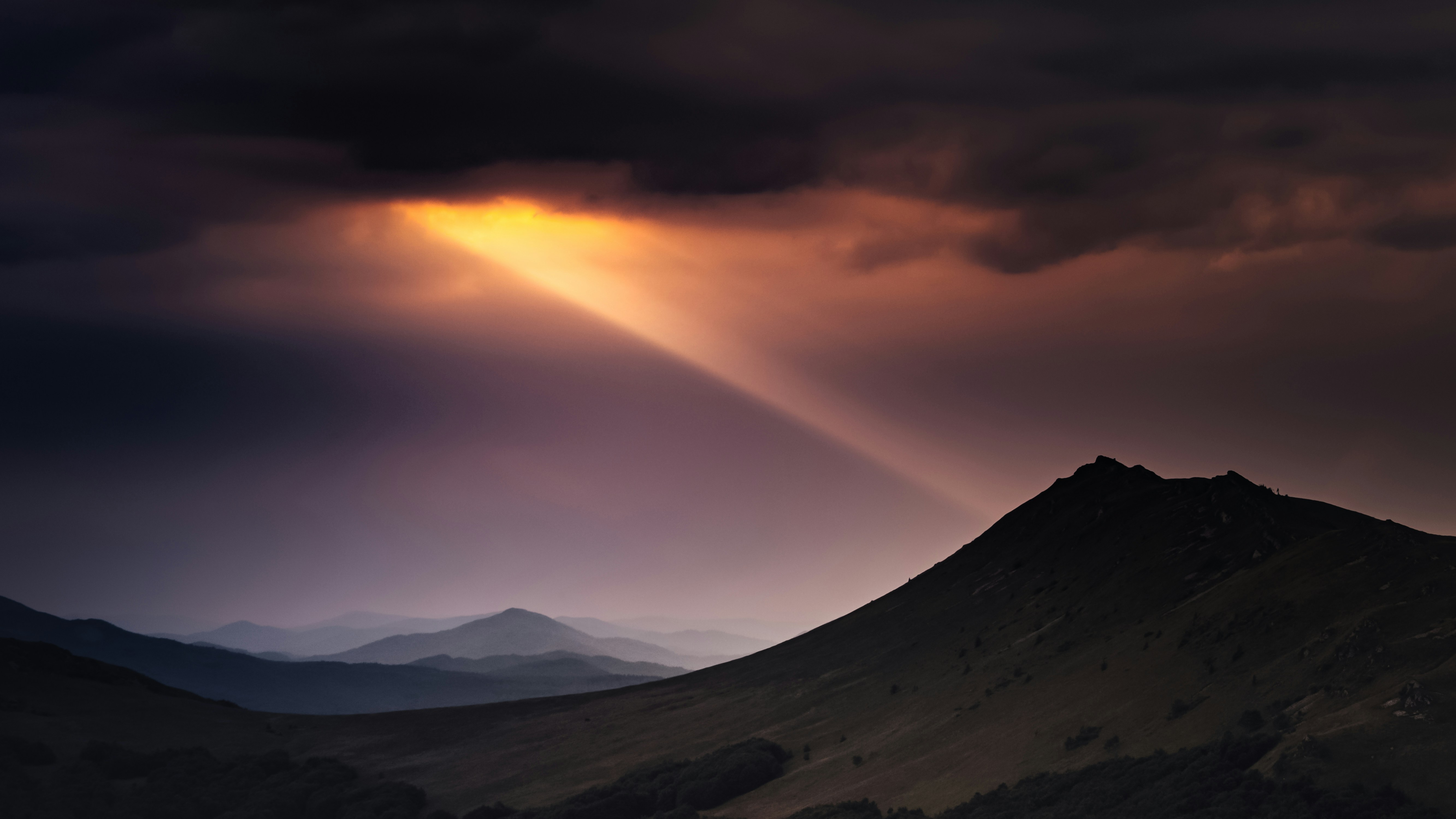 a dramatic mountain landscape at either dawn or dusk. dark, moody clouds fill the majority of the sky, pierced by a single, powerful ray of sunlight. this light illuminates the peaks of distant mountains and casts a golden glow on a grassy hillside in the foreground. the scene evokes a sense of awe and tranquility, showcasing the beauty of nature's contrasting elements.