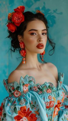 a woman with dark hair in a bun adorned with red roses wears dangling red earrings and an offtheshoulder aqua blue dress with red, orange and yellow embroidered flowers.  she is posed against a turquoise background.