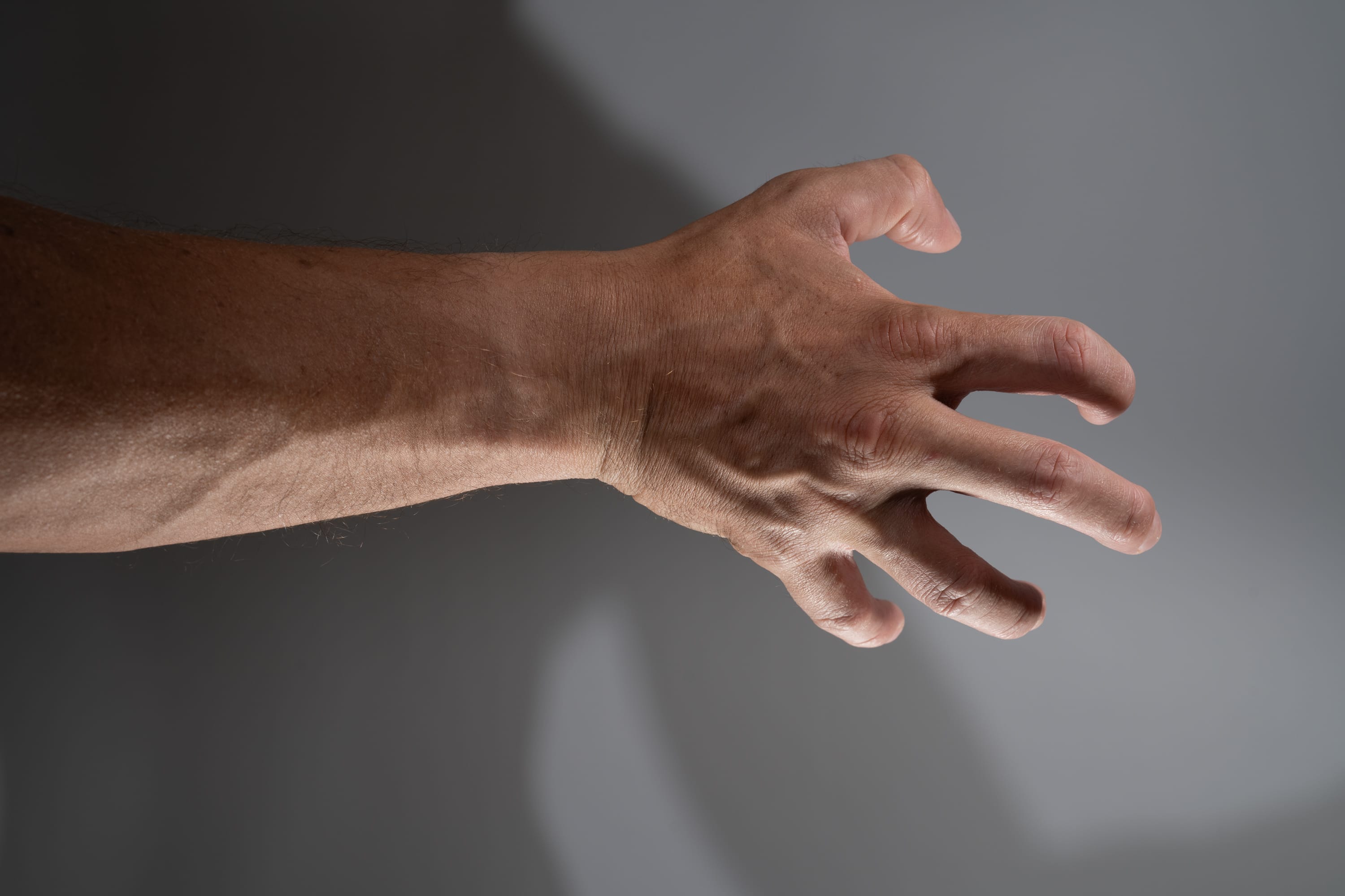 a closeup side view of a man's right arm and hand extended outward with fingers slightly curled.  the image is lit with natural light creating a shadow of the arm and hand on the wall behind.