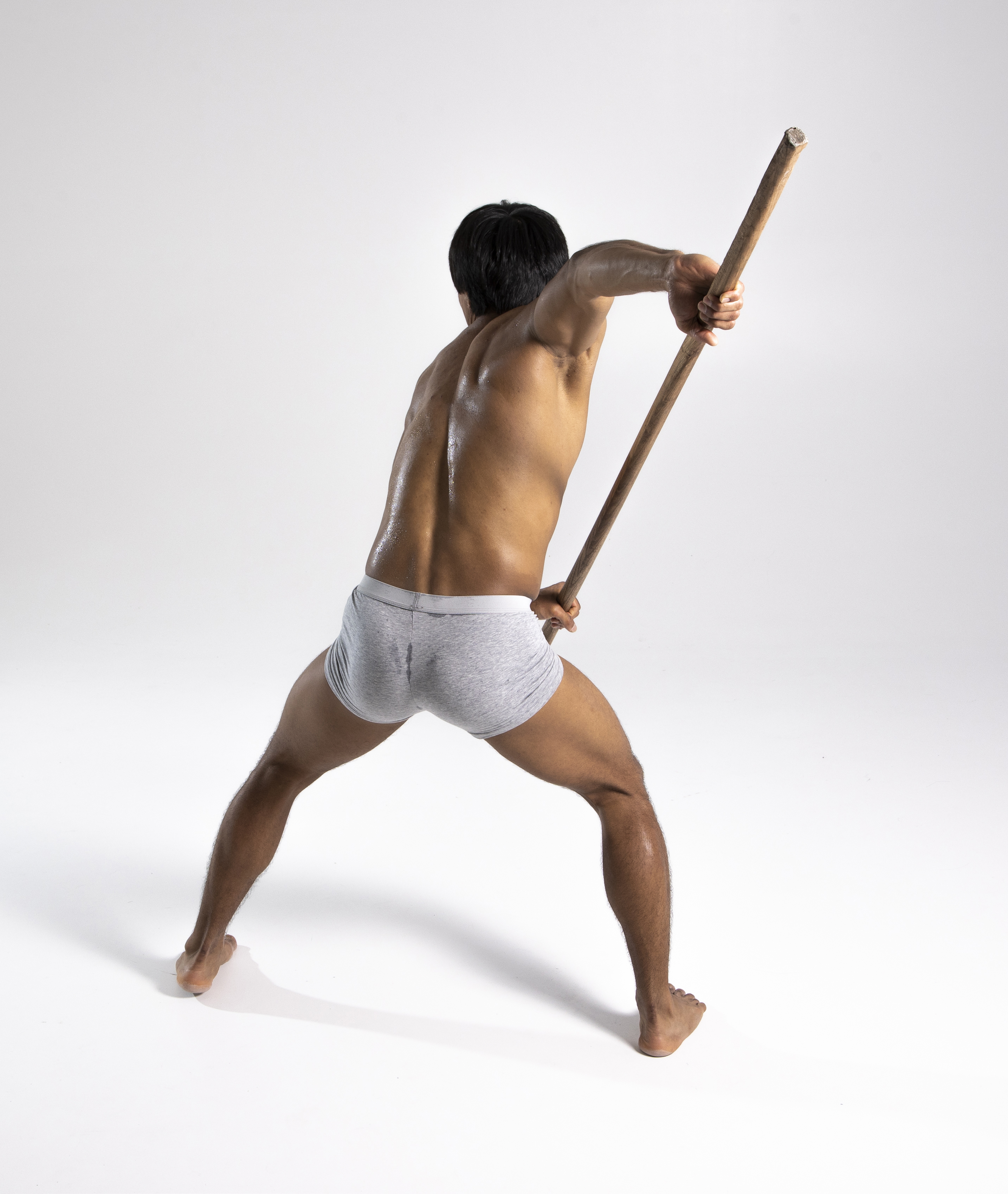 a rear view shot of an athletic asian man in gray underwear holding a wooden stick. he is barefoot and barechested, captured in a studio shot with a white background.