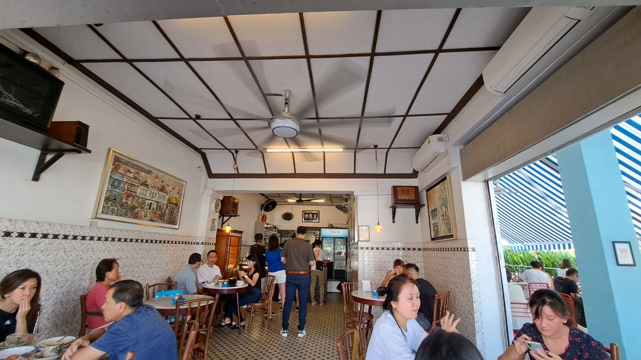 the image shows a cafe or restaurant interior with people seated at tables and enjoying their meals. the cafe has a traditional, vintage feel, with wooden tables and chairs, tiled floors, and a ceiling fan. there are people sitting at tables, chatting, and eating. the cafe has a window with a striped awning, which suggests it is a streetside location. the scene is bright and colorful, suggesting a lively atmosphere. the image captures the essence of a typical asian cafe or restaurant, where people come to socialize and enjoy good food.