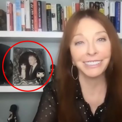 a woman with long dark hair is smiling and looking at the camera. she is wearing a black and white patterned top. there is a picture frame on the shelf behind her. the picture in the frame is of a woman with dark hair and a man with light hair.