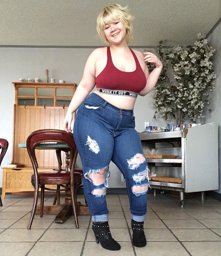 a blonde woman in a red crop top and ripped jeans is standing in a cafe, smiling at the camera. she is wearing ankle boots and has a confident expression on her face. the background is a simple cafe setting with tables and chairs.