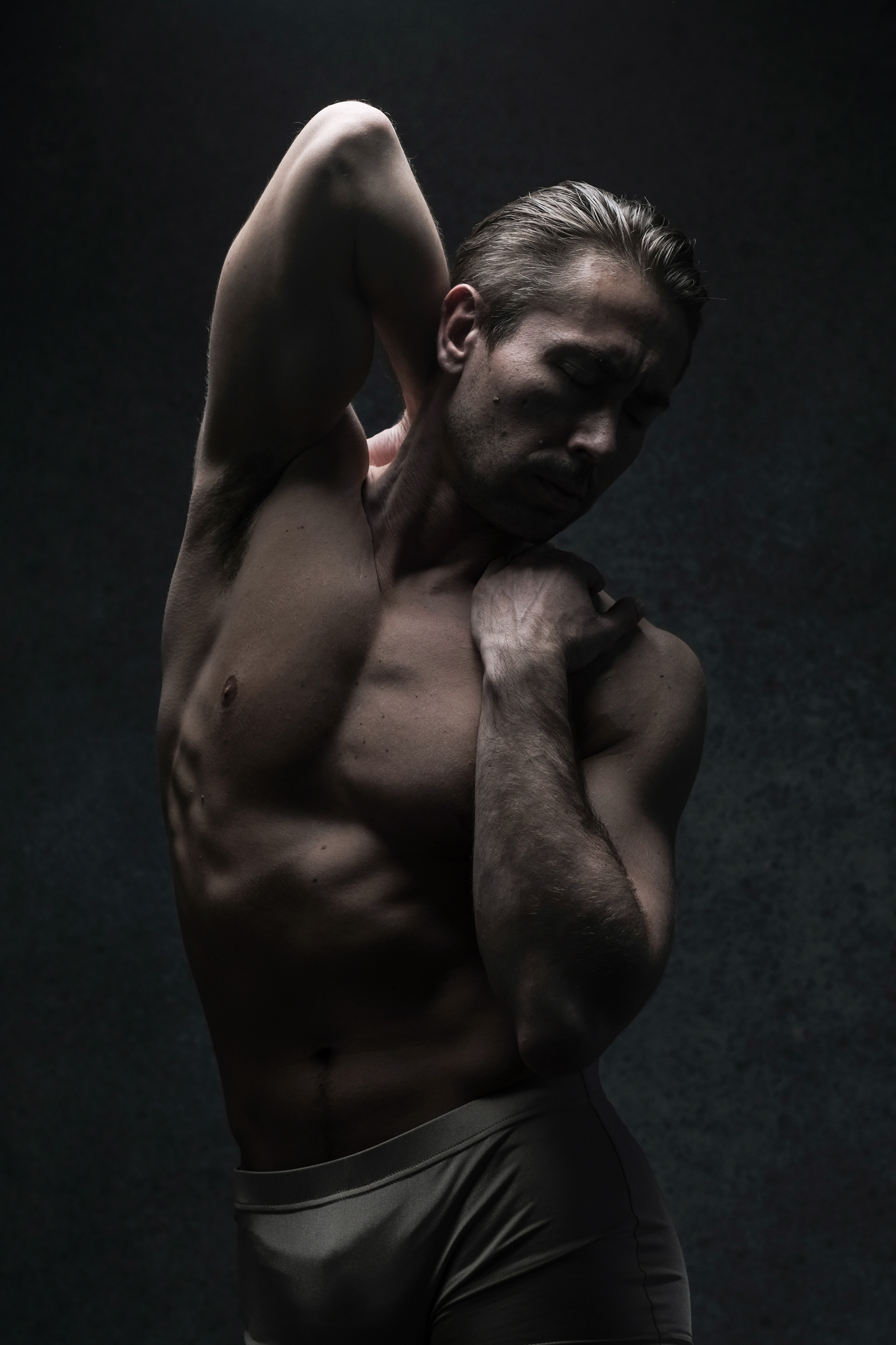 a muscular man in underwear is posing against a dark gray background. his eyes are closed and he is holding his arm behind his neck. the lighting is dramatic, creating a chiaroscuro effect.