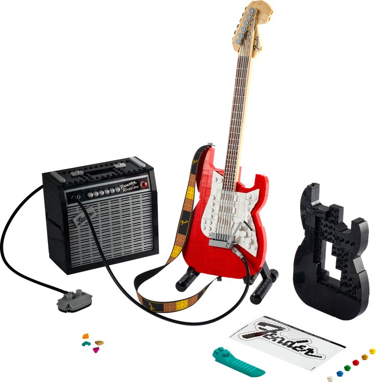 the image shows a lego ideas fender stratocaster set. the set includes a red and white lego guitar on a stand, a black lego amplifier with a power cable and foot pedal, a black lego guitar body, a guitar strap, a sticker with the fender logo, a green guitar pick, and a few small lego pieces. the set is placed on a white background.