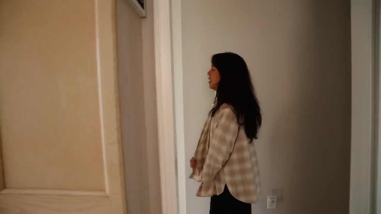 a young woman with long dark hair is standing in a hallway. she is wearing a beige and white plaid shirt and black pants. the walls are painted a light gray, and the lighting is soft and natural. the overall atmosphere is relaxed and casual.
