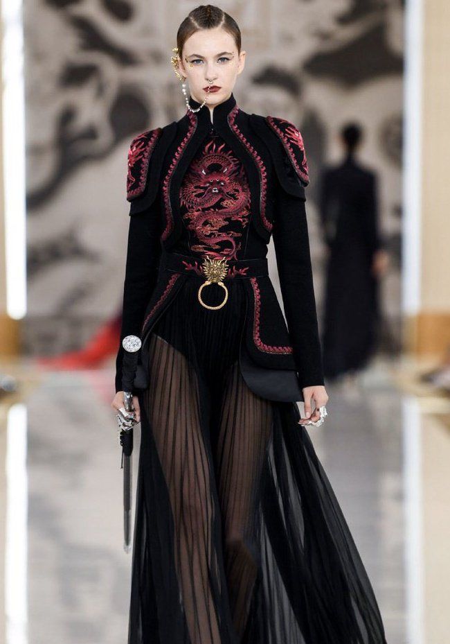 a model is walking on a runway wearing a black, orientalstyle dress with red dragon embroidery. the dress has a high collar, shoulder pads, a ring belt with a dragon clasp, and a sheer skirt. the model is also wearing a sword, an ear cuff, and a nose ring.