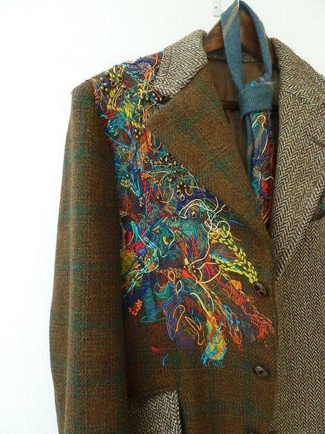 a closeup shot of a brown tweed jacket with a colorful, intricate embroidery design on the left shoulder. the embroidery consists of various threads and stitching creating a unique and artistic pattern. the jacket also features a brown button and pocket. a blue tie is visible on the jacket's collar. the fabric is a mix of brown and green tweed with a subtle herringbone pattern. the jacket is a unique and stylish piece of clothing that embodies the spirit of handcrafted art and fashion.