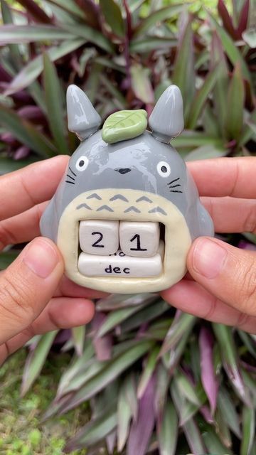 a pair of hands holds a totoroshaped perpetual calendar showing the date december 21st. the calendar is made of ceramic and has a green leaf on its head. the number blocks are white with black numbers and letters. the background is a lush green with a plant having purplegreen leaves.
