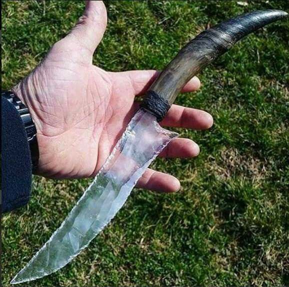 a person is holding an obsidian or glass knife with a darkcolored horn handle. the knife blade is transparent and very sharp looking. the background is a grassy lawn. the hand has a watch on the wrist. the knife appears to be handmade and unique.