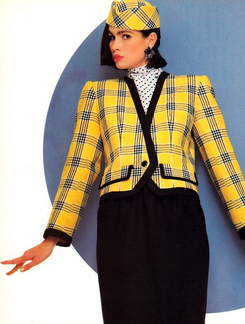 a woman is wearing a yellow plaid suit with a matching hat. the suit consists of a blazer with black trim and a black skirt. she also wears a polka dot turtleneck and black starshaped earrings. the background is a blue circle.