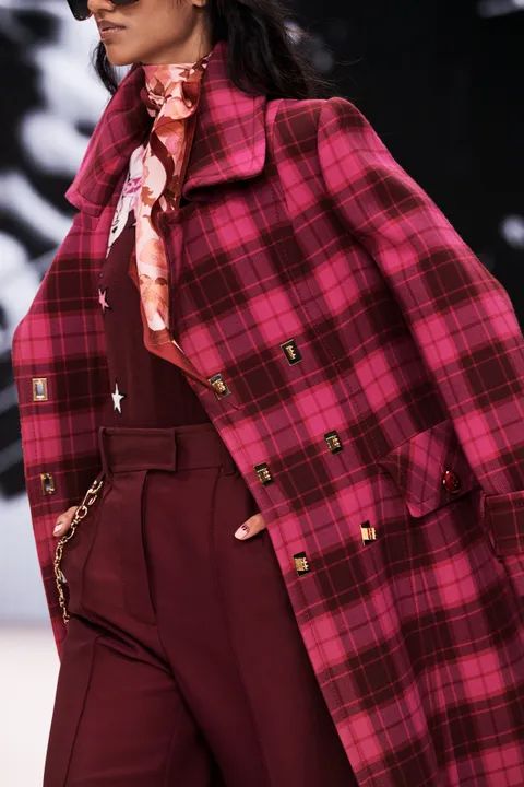 the image showcases a fashion model on a runway, wearing a striking long, doublebreasted coat.  the coat is a vibrant pink and burgundy plaid pattern. underneath, she wears a maroon colored dress and matching trousers.  she's accessorized with sunglasses, a patterned silk scarf at the neck, and a gold chain necklace that drapes partially down her coat. the overall aesthetic is elegant and fashionable, suggesting an autumn or winter collection.  the background is blurred, focusing attention on the model and her clothing.