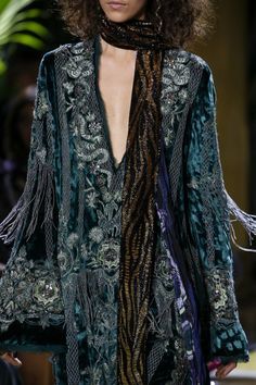 loseup view of a female model showcasing a teal velvet jacket or coat adorned with intricate silver embroidery. the jacket has long sleeves with fringe detailing at the edges. the model is wearing a long, dark brown and black striped scarf draped around her neck. her hair is dark brown and styled in loose waves. the background is blurry, suggesting a fashion runway or photoshoot setting.  the overall aesthetic is bohemian and luxurious.