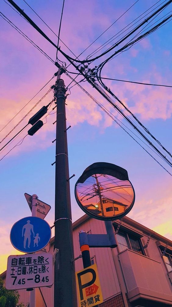 the image is an aesthetic street photography shot taken from a low angle. the focal point is the intersection of wires and a streetlight pole set against a beautiful sunset sky. the sky exhibits a gradient of pink, purple, and blue hues. a convex mirror is attached to the pole, reflecting the buildings and the street below. adding to the urban ambiance, there's a "no bicycle parking" sign and a pedestrian crosswalk sign on the left. the overall composition exudes a sense of calm and the warm tones of the sunset enhance the visual appeal.