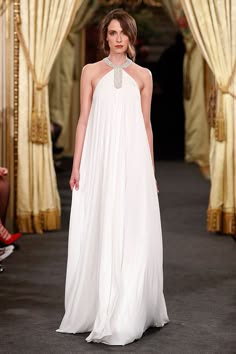 a woman in a long white dress stands on a runway, posing for the camera. the dress is sleeveless and has a flowy, silky fabric. the woman's hair is pulled back and she is wearing minimal makeup.  the background is a luxurious, goldenhued setting, suggesting a highfashion event. the dress's style is elegant and timeless, suitable for a wedding or other formal occasions.  the model's confidence and poise are accentuated by the dress's simplicity and flow.