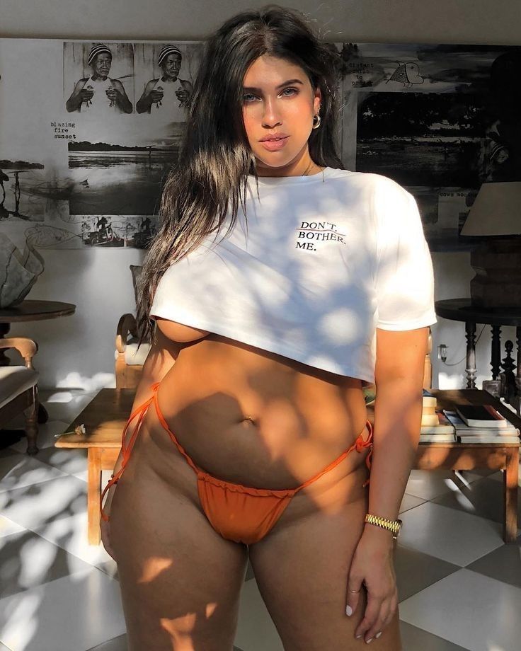 a woman is posing in a crop top and bikini bottoms. she is wearing a white crop top tshirt with the words "don't bother me." written on it. the bottoms are orange bikini bottoms. she is wearing a watch on her left wrist and what appear to be hoop earrings. her hair is brown and shoulderlength. she is in front of a wall with various artworks on it. there is a table with books on it and a chair next to her. the floor is checkered.