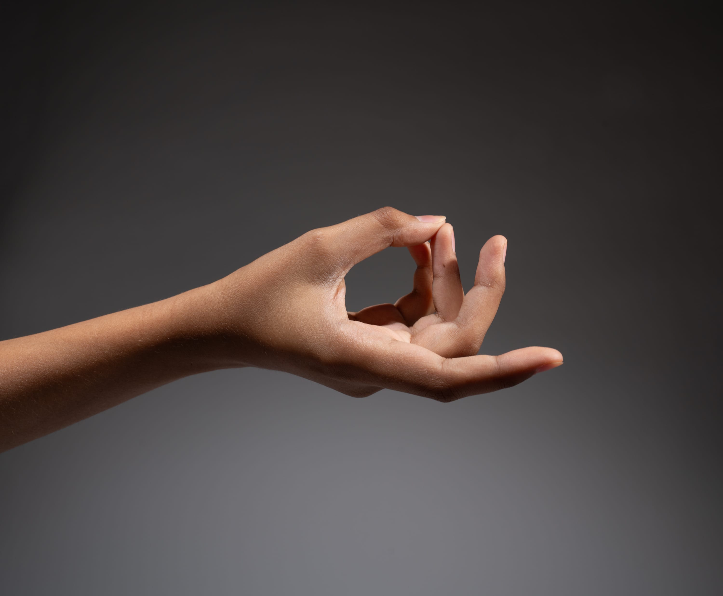 a woman's hand is making a gyan mudra gesture, which is often used in meditation and yoga. the hand is positioned against a gray background, with studio lighting highlighting the gesture.