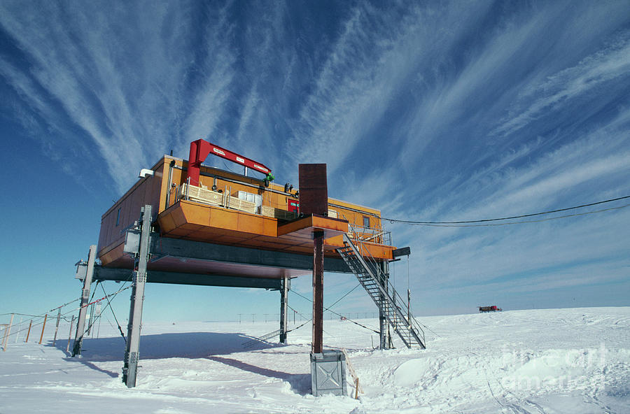 a yellow building on stilts sits on a snowy landscape in antarctica. the building is part of a research station, and the sky is filled with wispy white clouds against a blue backdrop.  a small red vehicle is visible in the distance on the snowy horizon.