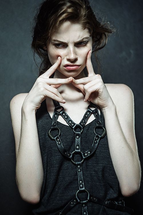 a young woman with brunette hair is dressed in black leather clothing. she is wearing a harness over a black tank top, and her face is serious and thoughtful. she is looking down with her fingers on her cheeks. the lighting is dramatic and creates a strong contrast between the woman and the dark background.