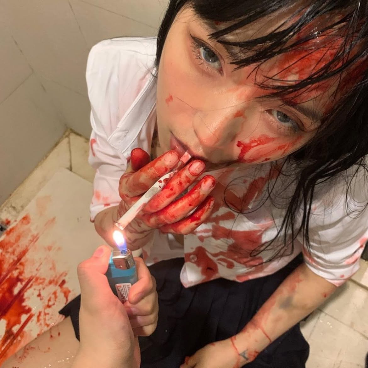 a woman in a school uniform is covered in blood and holding a cigarette in her mouth. another hand holds a lighter to the cigarette. she has bruises on her arm. there's blood splattered on the wall and floor behind her.