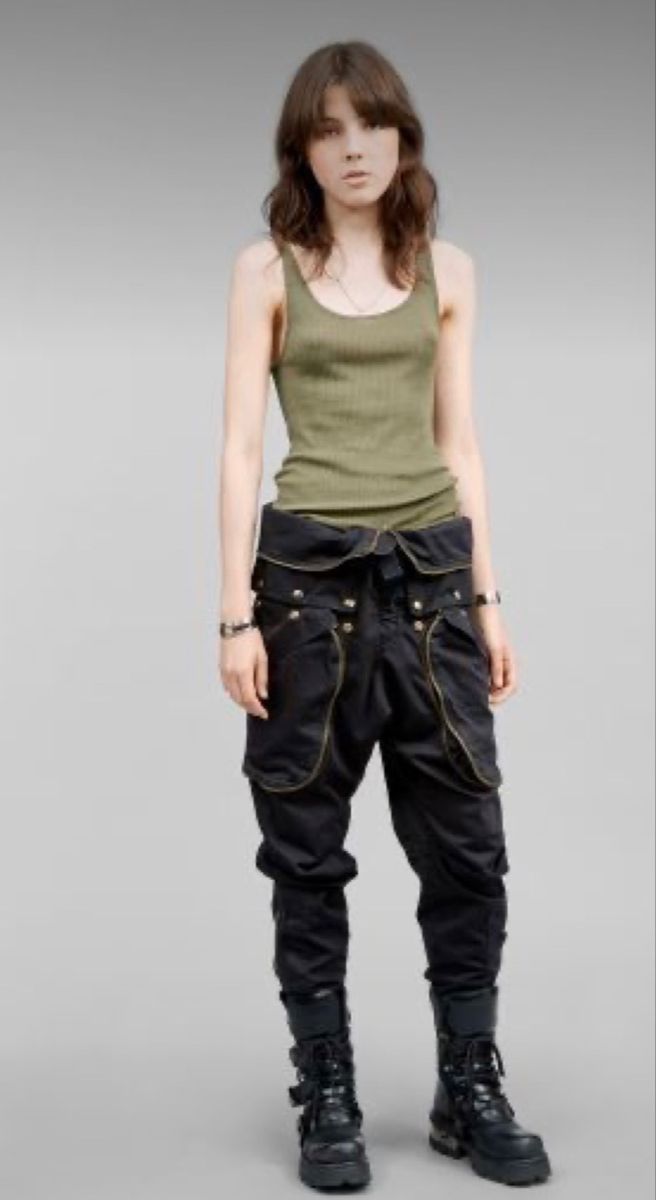 a fullbody shot of a female model standing against a light grey background. she is wearing a light olive green tank top and black cargo pants with a unique design around the waist and gold detailing. her dark brown hair is styled loosely around her shoulders. she is wearing black combat boots with buckles. the overall style is edgy and slightly militaryinspired. the lighting is even and wellbalanced.