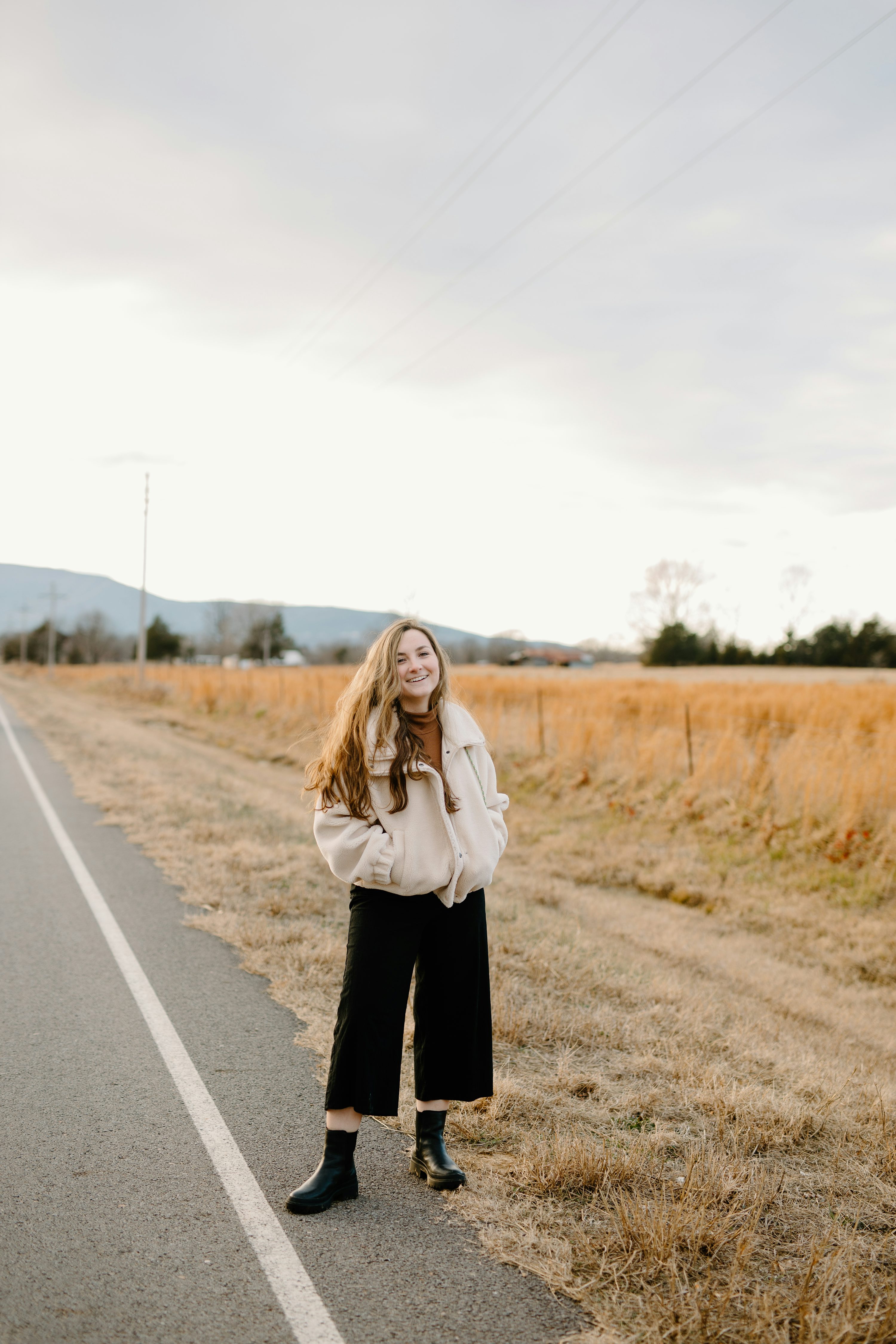 a young woman stands on the side of a country road, smiling at the camera. she is wearing a creamcolored jacket, black culottes, and black ankle boots. the road is asphalt and has a white line down the middle.  the background is a field of dry, golden grass and distant mountains under a cloudy sky. the overall mood is peaceful and serene. the lighting suggests it is likely late afternoon or early evening.