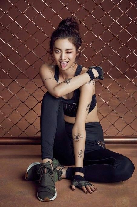 a young asian woman with a sporty look poses in a studio in front of a chainlink fence.  she's wearing black leggings and sneakers and has tattoos on her arms.  she's sitting with one leg bent and the other straight out, holding a pair of black gloves in her hand.  she's looking at the camera and sticking her tongue out in a playful manner.