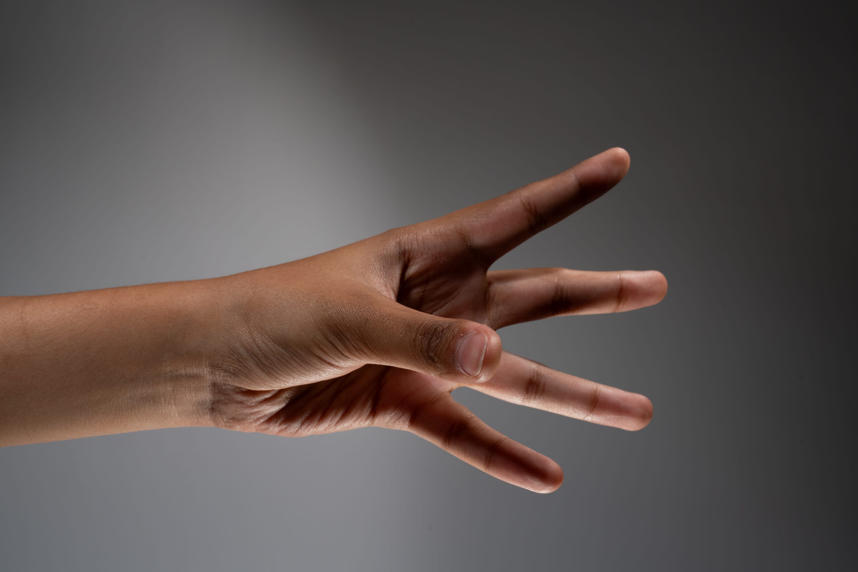 an image of a hand with dark skin tone against a gray background, illuminated by a spotlight. the hand is extending three fingers, indicating the number three.