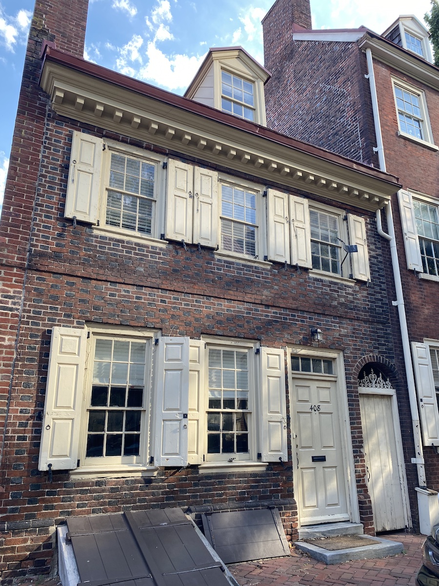 a threestory brick building with creamcolored window shutters, door, and trim. the building has a dormer window on the roof and a chimney on the left side. there is a small door next to the main door and basement doors visible near the ground. the building is made of dark red bricks.
