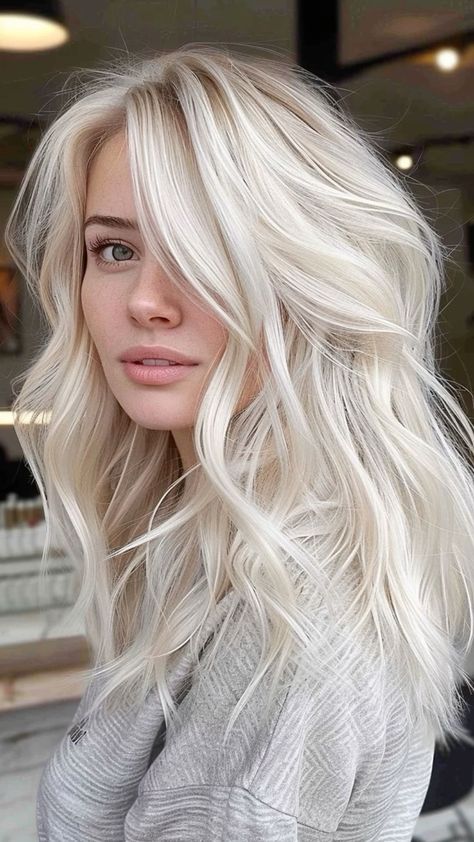 a woman with long, wavy platinum blonde hair is looking offcamera. she is wearing a gray sweater and the background is blurred. the lighting is soft and natural.
