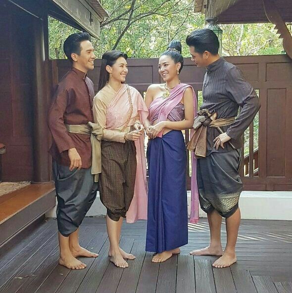 a group of four friends, two men and two women, are standing on a wooden floor, wearing traditional thai clothing. they are smiling and looking at each other, enjoying their time together. the men are wearing longsleeved shirts with pants and sashes, while the women are wearing long dresses with colorful decorations. the background is a wooden building, likely a temple or home. this image captures the beauty of traditional thai clothing and the joyful camaraderie of friends.