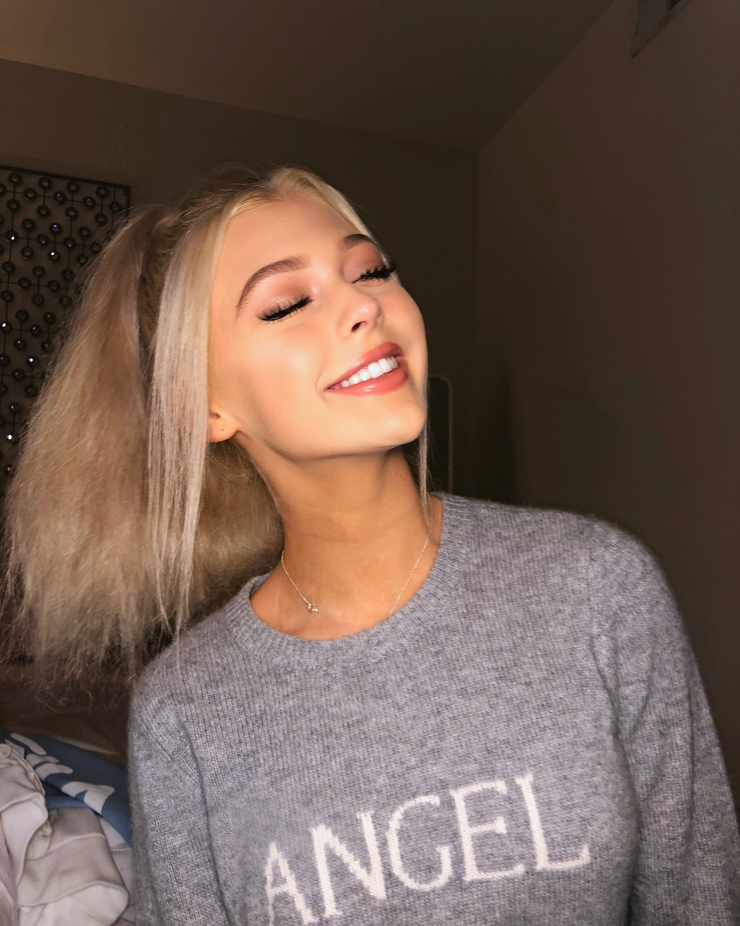 a young blonde woman smiles with her eyes closed, wearing a grey sweater with the word "angel" written in white. she also wears a delicate silver necklace. the photo appears to be taken indoors in a room with a dark background. her blonde hair is styled in a half ponytail.