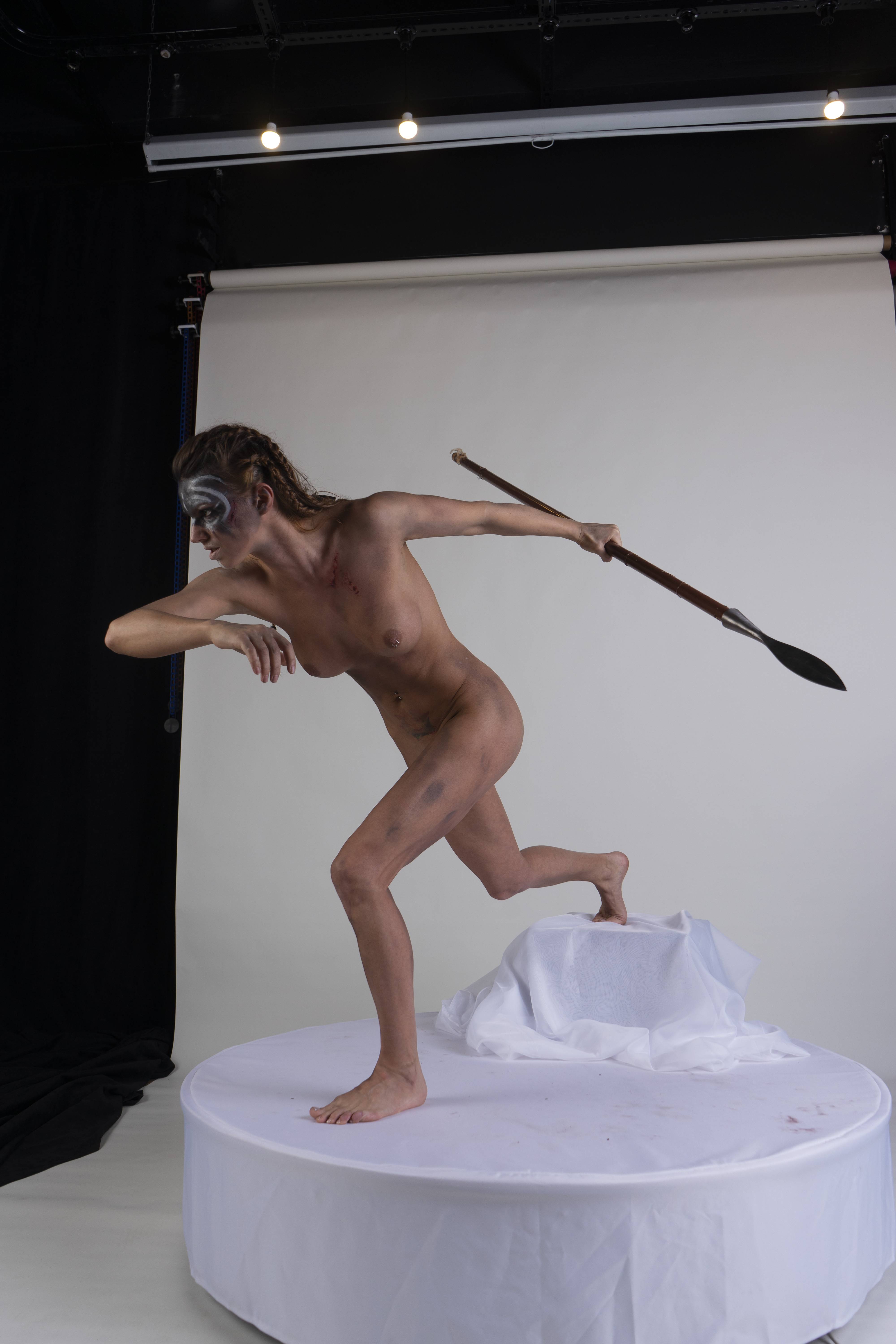 a nude woman in warrior makeup and braids is running across a white pedestal, holding a spear. she is in a studio with a white background.