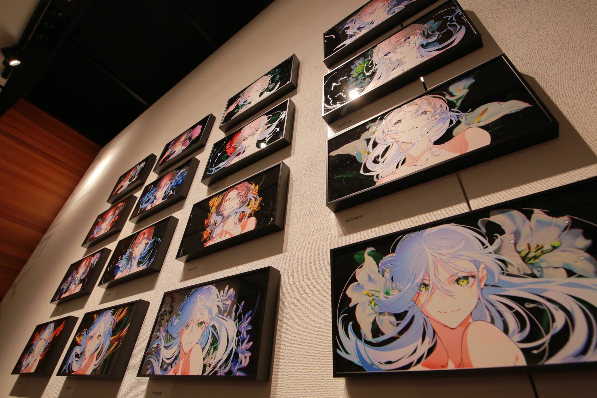 the image showcases a collection of framed animestyle artwork displayed on a white wall. each piece features a female character with flowing blue hair, set against a backdrop of vibrant, abstract designs and floral elements. the artwork is illuminated by artificial lighting, creating a dramatic and captivating ambiance. the lowangle perspective emphasizes the scale of the display and draws the viewer's eye upwards, enhancing the sense of immersion in the artistic space.