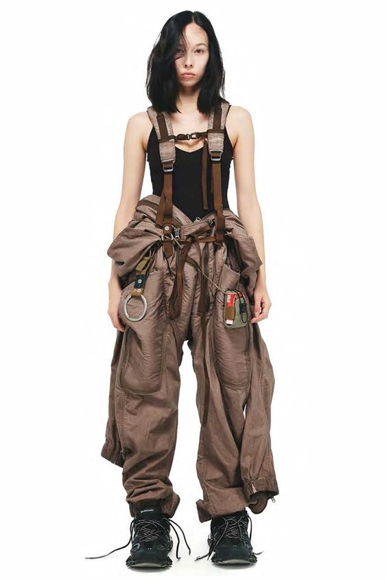 a young woman is standing in front of a white background wearing a brown jumpsuit with black straps. the straps have multiple buckles and metal attachments, some dangling loosely. she is wearing black shoes and her arms are at her side. her hair is black and she has a slightly serious expression on her face.