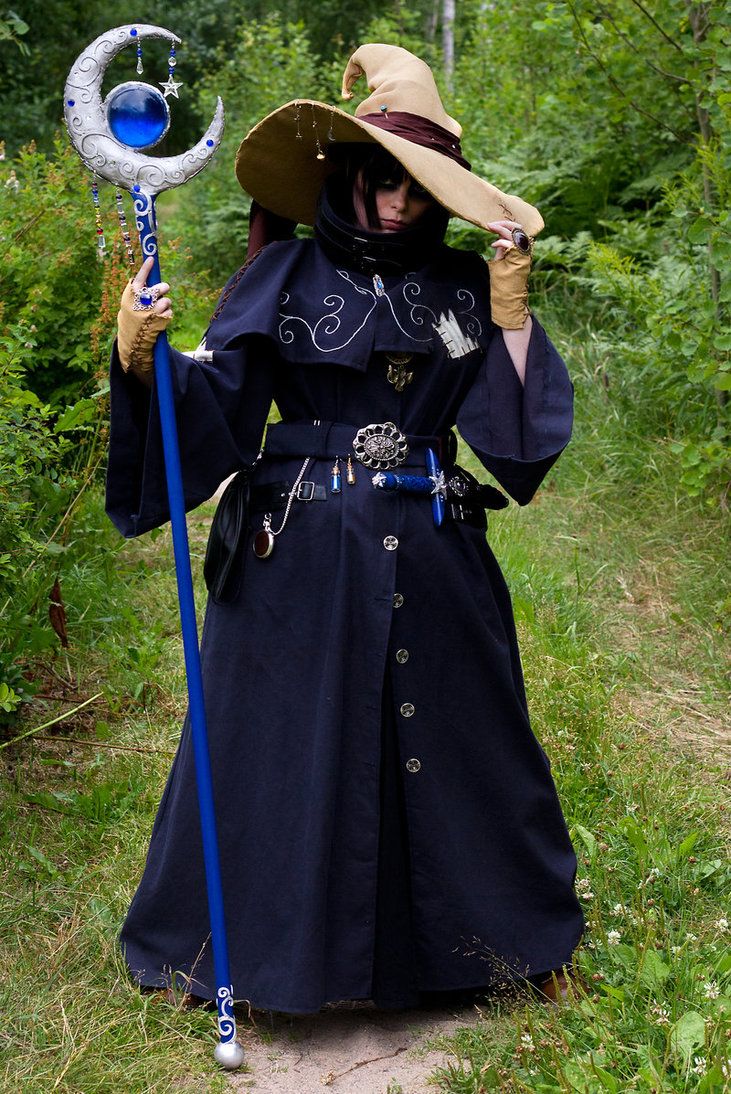 a woman in a wizard costume is holding a staff in her left hand and touching her hat with her right hand. she is dressed in a long blue robe, with a widebrimmed hat, and wearing gloves. the costume has various accessories attached to the belt. she is standing on a trail in a grassy area, with trees and bushes in the background. the staff is silver and blue, with a crescent moon shape at the top, containing a blue orb. the background is blurred.