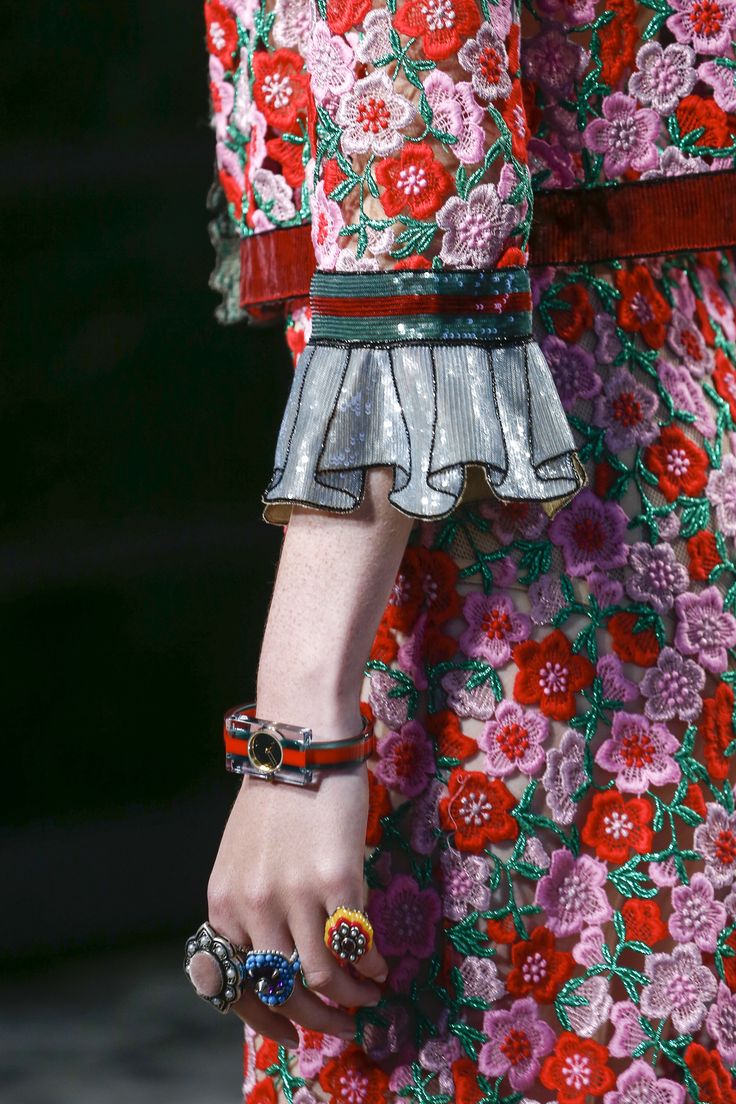 the image is a closeup shot focusing on the intricate details of a gucci dress featured on a runway model. the dress is predominantly red and pink, showcasing a delicate embroidered floral lace pattern with added sequins and beads.  the sleeve features a ruffled detail with a silver sequin trim.  the model's hand is visible, adorned with several colorful rings and a unique, clearfaced wristwatch with red and green detailing, matching the color scheme of the dress. the overall aesthetic is luxurious and highly detailed, showcasing the craftsmanship of the garment and its accessories.