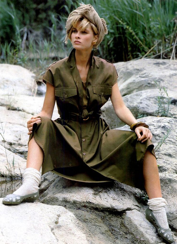 a highfashion photograph from the 1980s features a blonde female model, casually sitting on rocks in a natural outdoor setting.  she is wearing a loosefitting, olivegreen safaristyle dress,  a light beige head scarf,  lightcolored socks and clogs. her pose is relaxed and her expression is somewhat pensive. the background includes lush greenery and rocky terrain, suggesting a wilderness or travel theme. the overall aesthetic is indicative of high fashion photography from that era.