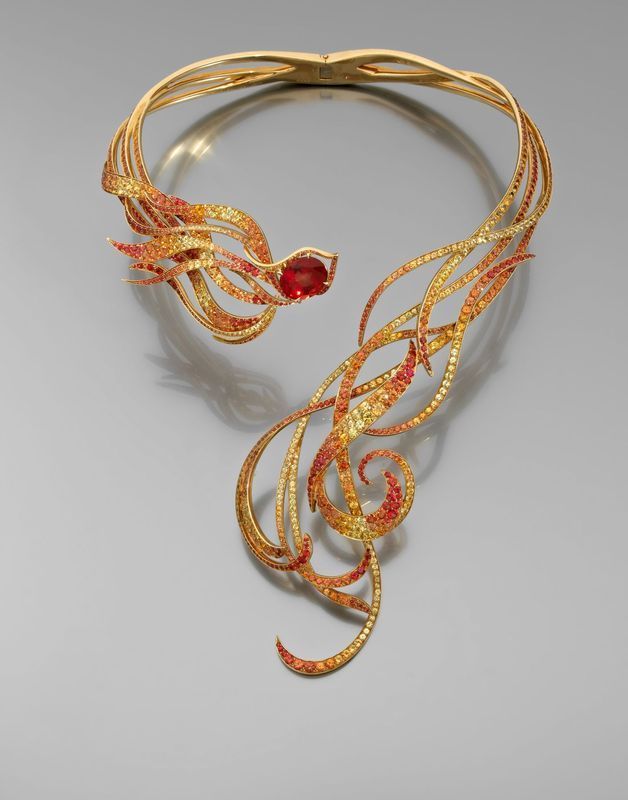 a gold necklace featuring a stylized fire or flame design with red, orange, and yellow gemstones arranged on swirling, fluid lines. the central focus is a large red gem at one end, suggesting a head or focal point for the fiery design. the necklace appears ornate and luxurious.