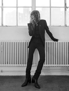 a woman in a pinstripe suit and boots standing in a studio. the woman is wearing a cigarette in her mouth and looks serious. she is posed against a wall with a radiator in the background.