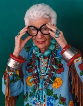 the image is a closeup portrait of iris apfel, a renowned fashion icon. she is an older woman with short, white hair, wearing large round glasses. apfel is adorned with an abundance of turquoise and other colorful native americaninspired jewelry, including a substantial beaded necklace, numerous bracelets and rings. her clothing is a light blue, embroidered blouse with floral designs and long fringes. her hands are gently adjusting her glasses, her expression is calm and selfassured. the background is a simple, solid teal. the overall impression is one of vibrant style and personal expression.