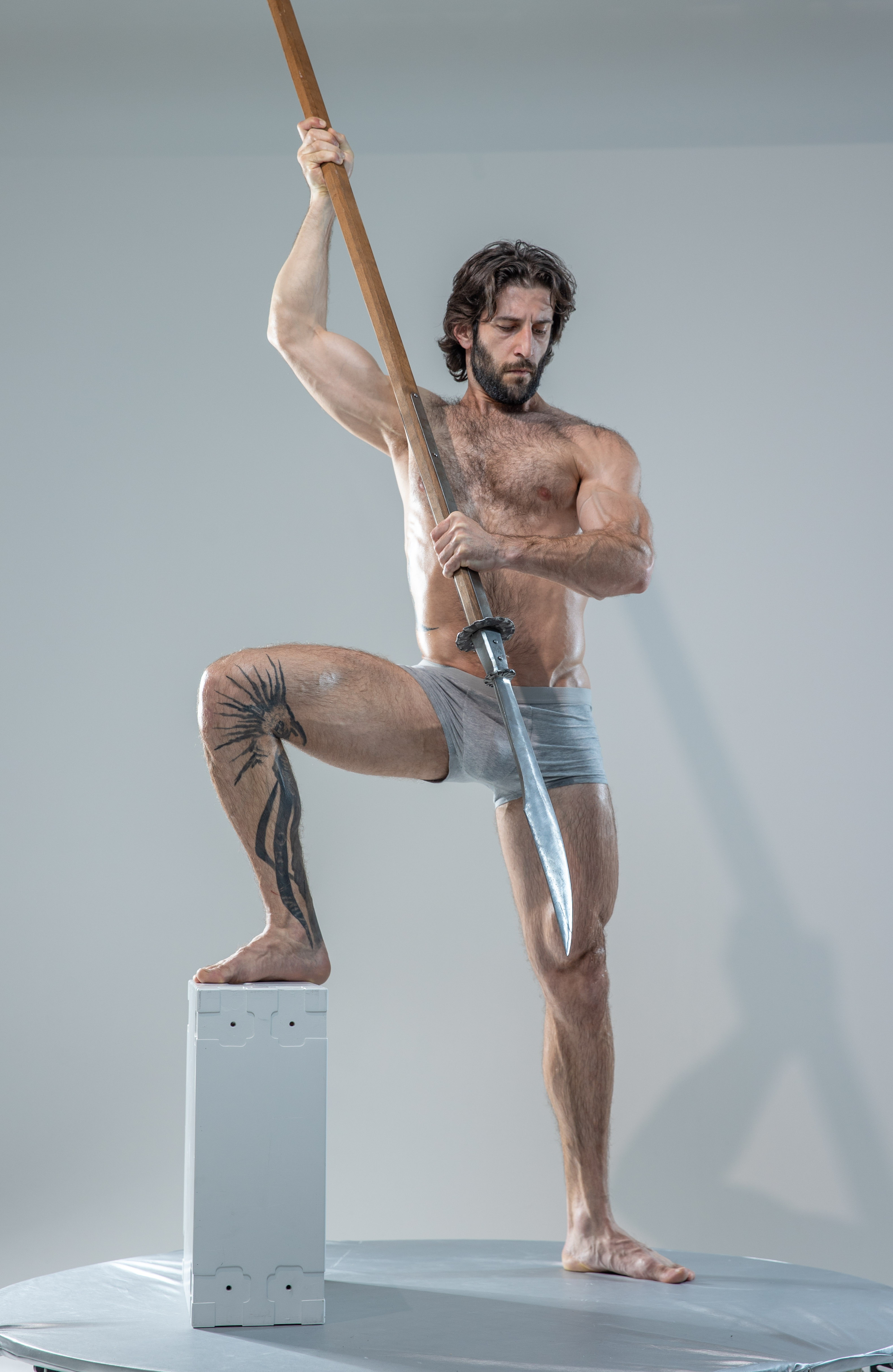 a muscular, bearded man with long hair is posed in a photography studio. he stands on a white platform and holds a spear with a determined expression. the man wears only gray underwear, showcasing his physique and a tattoo on his leg. the studio lighting casts soft shadows, highlighting his muscles and the spear.