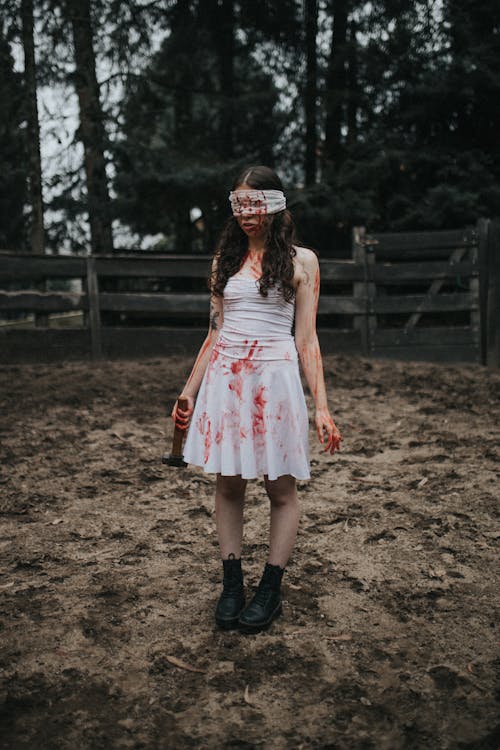 the image shows a young woman with long brown hair, blindfolded with a white cloth, wearing a white dress splattered with what appears to be blood. she holds an axe in her left hand.  her attire and the bloodstains suggest a horror or thriller theme. she's standing in a dirt area, possibly a farmyard, with a wooden fence visible in the background and a dark forest forming a backdrop. the overall mood is dark, mysterious, and unsettling.  the style is reminiscent of horror films or halloween imagery.