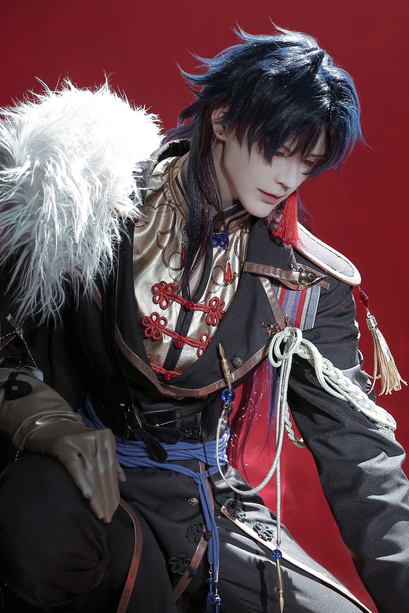 a young man is cosplaying as a character from an anime or manga. the character's name is unknown but they are wearing a traditional chinese costume with a black and red color scheme. the character's costume has a white furry collar, blue sashes, and a variety of other accessories. the man is posing with his head slightly bowed, as if they are thinking about something or experiencing a moment of sadness. they are looking down at something with their hands on their knees.  the background is a solid red color.