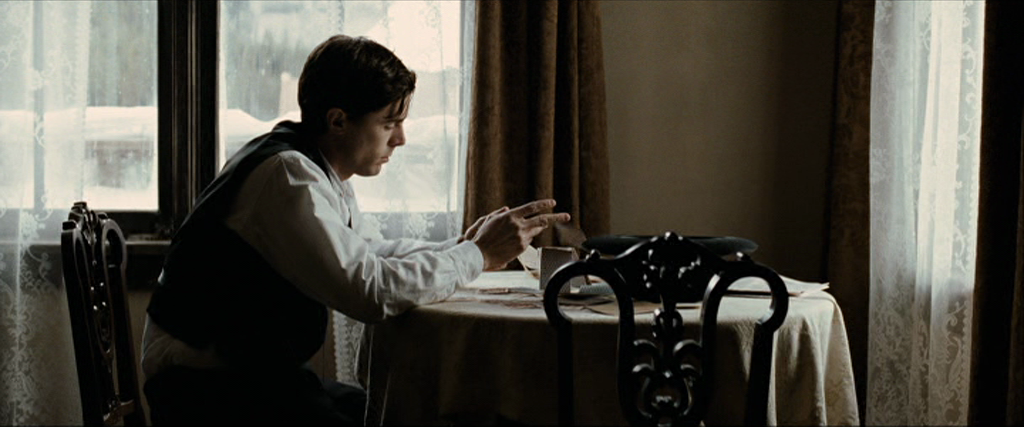 a man is sitting at a table in a dimly lit room. he is reading or writing something on a piece of paper. there is a window behind him and a chair to his right. he is dressed in a suit and a vest. he appears to be focused on his task. the image has a vintage feel to it.