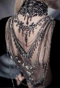 a closeup of a woman's neck adorned with several layers of elaborate silver necklaces. the necklaces are studded with rhinestones and pearls, giving the image a glamorous and elegant aesthetic. the woman's hair is styled in a sleek manner, and her skin has a glow, further enhancing the overall luxurious feel. the image captures the intricate details of the jewelry, showcasing the craftsmanship and opulence of the accessories.