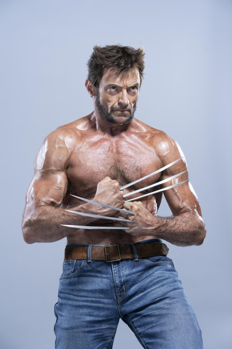 the image shows hugh jackman dressed as wolverine. he is shirtless, revealing his muscular physique, and wearing jeans with a brown belt. his signature claws are extended from his knuckles. he has a beard and a determined expression on his face. the background is a light gray or blue.